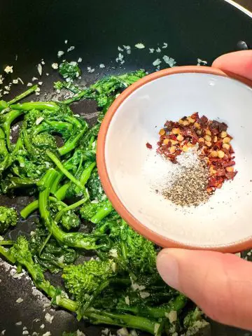 Sautéed Rapini, garlic, and oil in a pan with a white bowl containing red chili flakes, salt, and pepper being held by someone above the rapini in the pan.