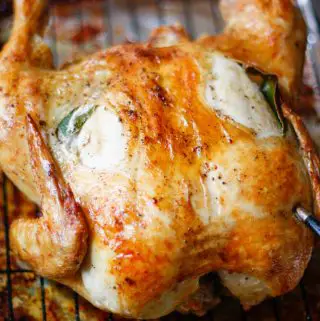 A roast chicken with crispy skin with holes in the skin that have been stuffed with bay leaves and garlic. The chicken is on a wire rack in a roasting tray and there is also garlic and a bay leaf in the cavity of the chicken and a MEATER thermometer is inserted in the chicken.
