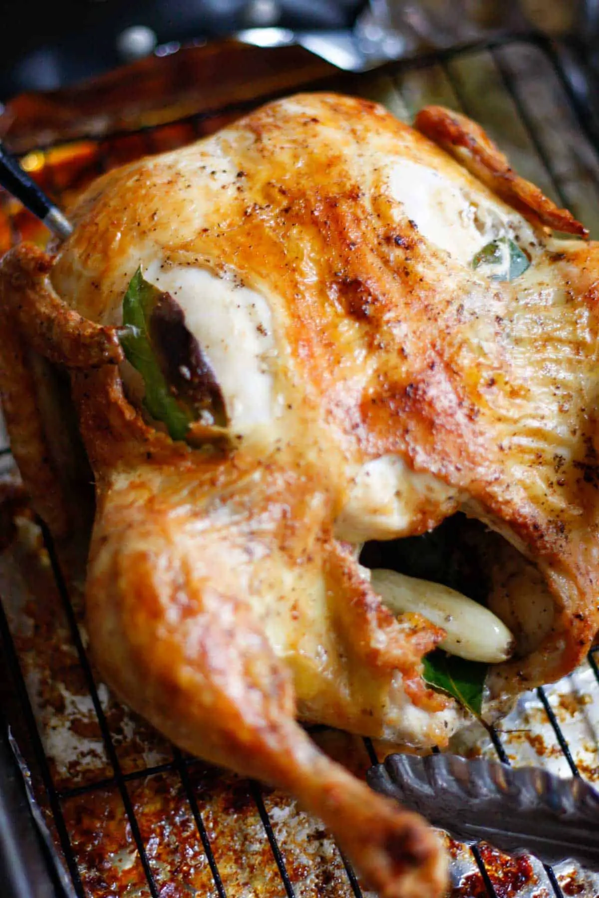 A roast chicken with crispy skin with holes in the skin that have been stuffed with bay leaves and garlic. The chicken is on a wire rack in a roasting tray and there is also garlic and a bay leaf in the cavity of the chicken.