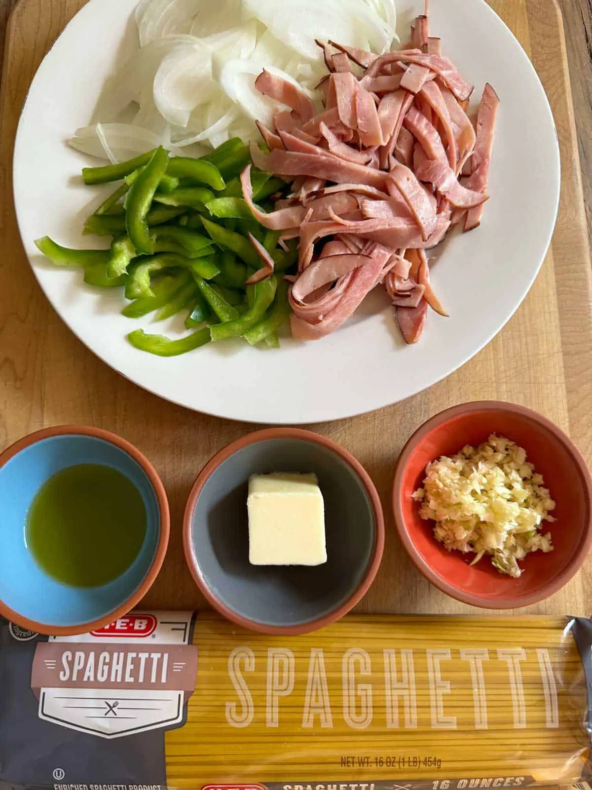 A white plate containing slices of ham, green bell pepper and onion. A bag of spaghetti and bowls containing olive oil, butter, and minced garlic.