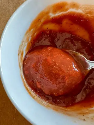 A ketchup based sauce in a white bowl. There is a spoon containing some of the sauce in the bowl.