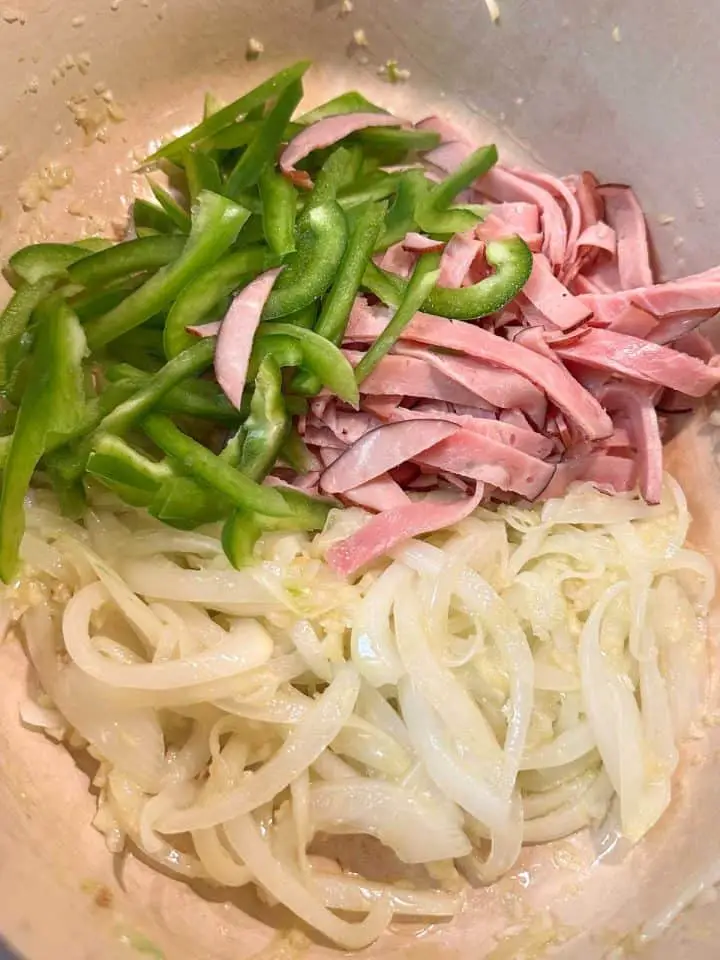 A dutch oven containing sliced ham, sliced green bell pepper and onions with minced garlic.