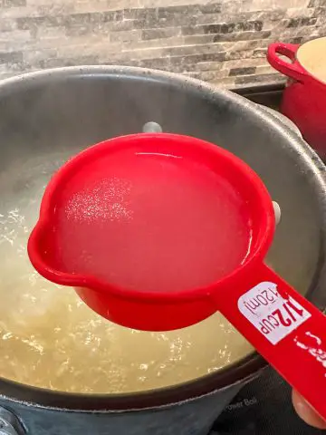 A large stock pot filled with boiling water and spaghetti with a red ½ cup measuring cup containing some of the pasta water poised above the stock pot. There is part of a red Dutch oven in the background.
