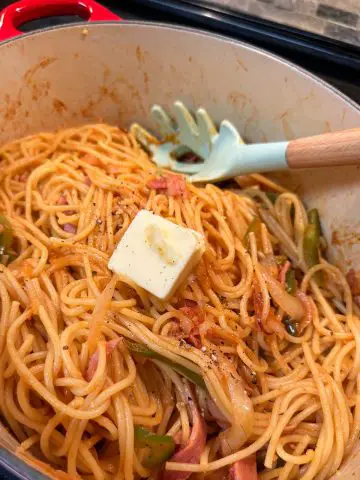 Japanese spaghetti with ketchup, slices of green bell pepper and onions in a dutch oven with a dab of butter and black pepper. There is a blue spaghetti server resting on the pasta.