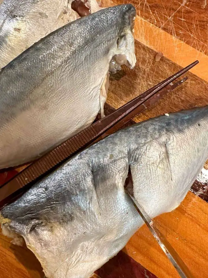 Uncooked mackerel on a cutting board. There is a knife opening a slit that has been cut into the flesh of one of the fish, and a pair of tweezers.