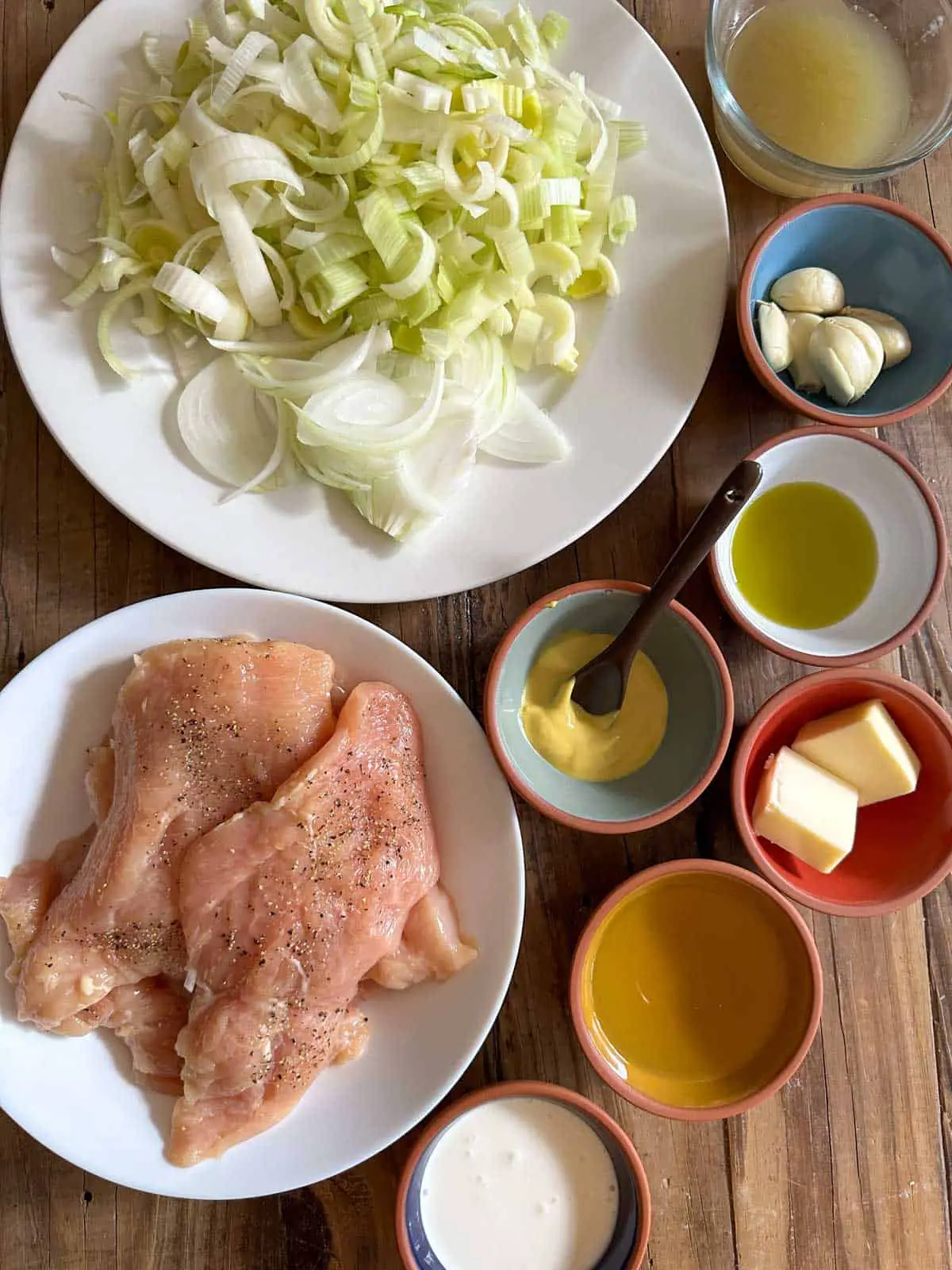 A white bowl filled with sliced leeks and onions, and another white bowl containing seasoned chicken breasts. Small bowls containing chicken broth, garlic, olive oil, mustard, butter, wine, and heavy cream.