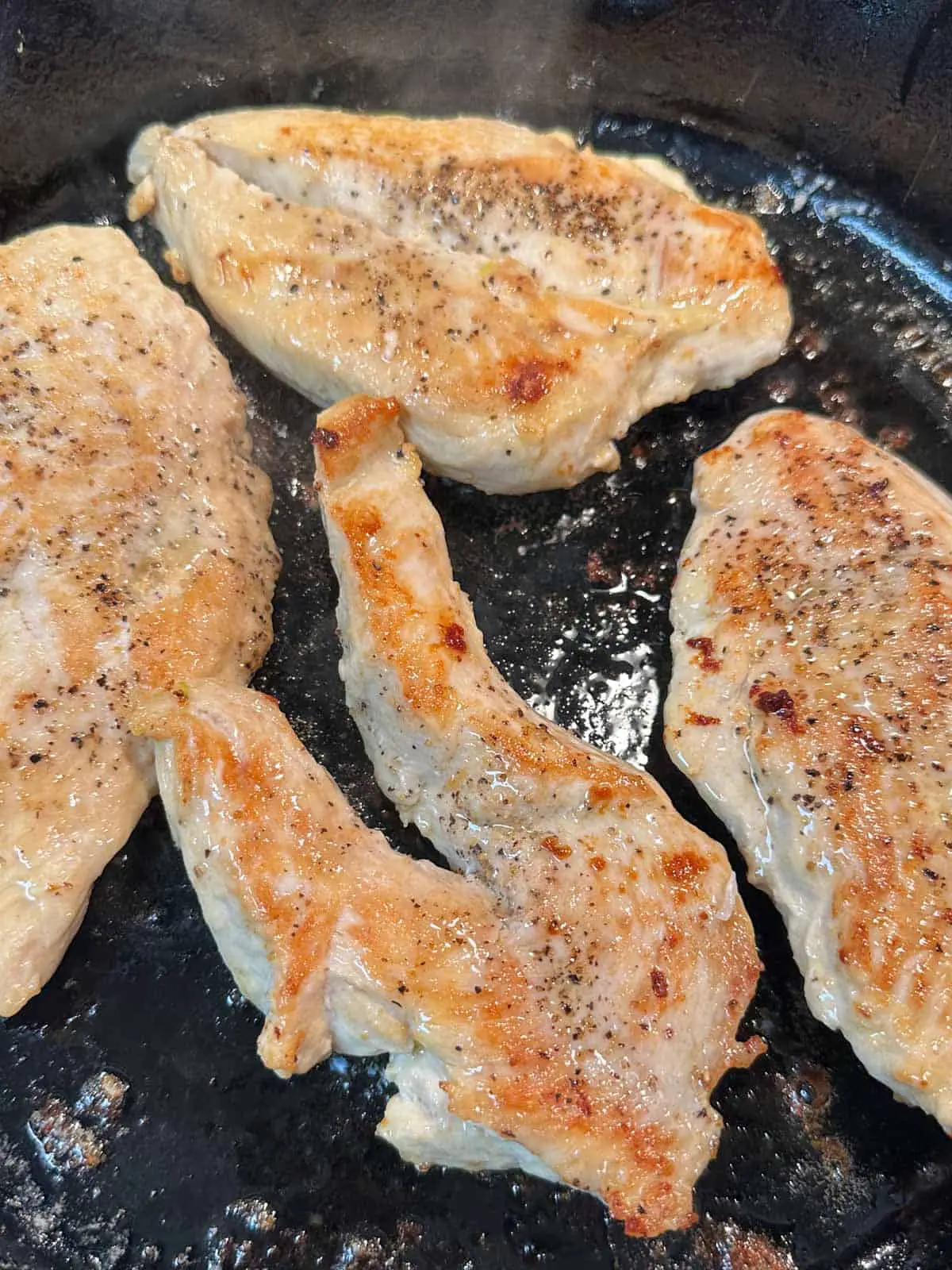 Browned pieces of chicken breast seasoned with pepper cooking in a cast iron pan.