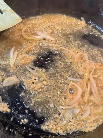 Sliced onion and minced garlic in melted butter and olive oil cooking in a cast iron pan. There is a blue silicone utensil in the top left of the picture resting on the pan.