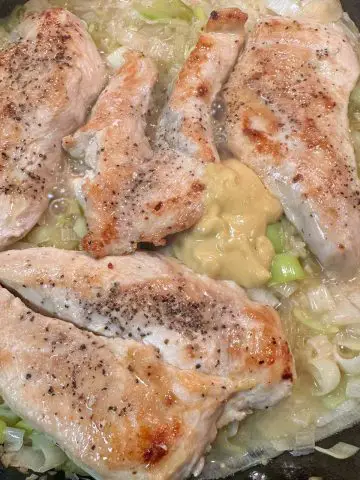 Chicken breast pieces cooking in broth in a cast iron pan with leeks and a dollop of mustard.