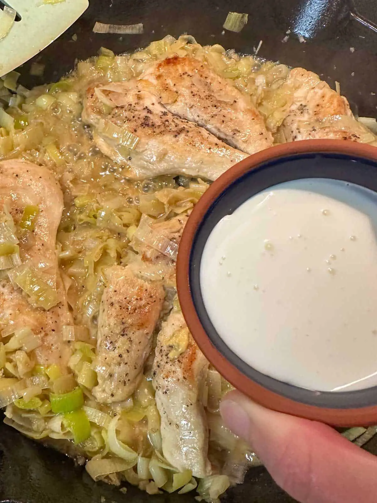 Chicken cooked with leeks in a cast iron pan. There is a blue silicone utensil on the top left and someone holding a small bowl with heavy cream poised over the chicken.
