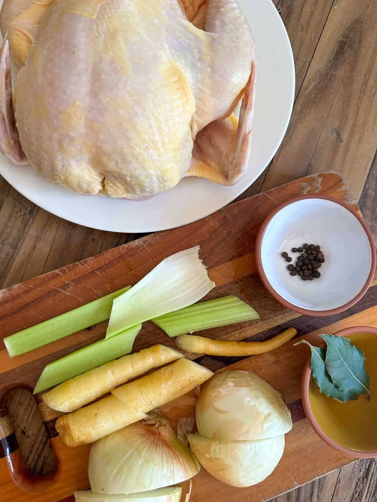 A whole uncooked chicken on a white plate. There is a wooden cutting board with celery, carrots, onions, bay leaves and peppercorns on it.