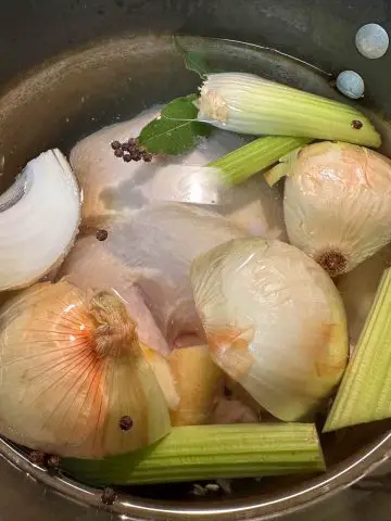 A large stockpot containing water and onions, celery, bay leaf, carrots, chicken, and peppercorns are visible.