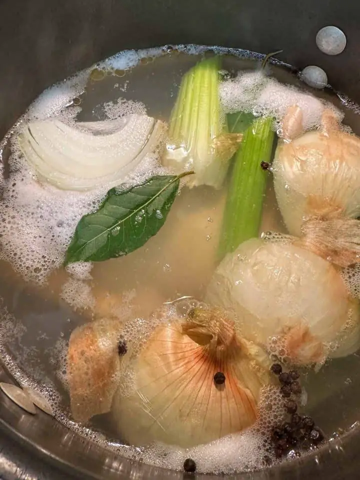 A large stockpot containing water and onions, celery, bay leaf, chicken, and peppercorns are visible. There is scum forming at the top.