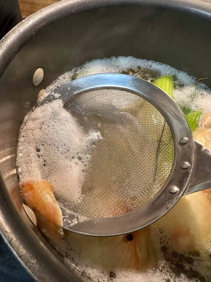 A large stockpot containing water and onions, celery, and peppercorns are visible. There is scum at the top and a strainer spoon is scooping up some of the scum.