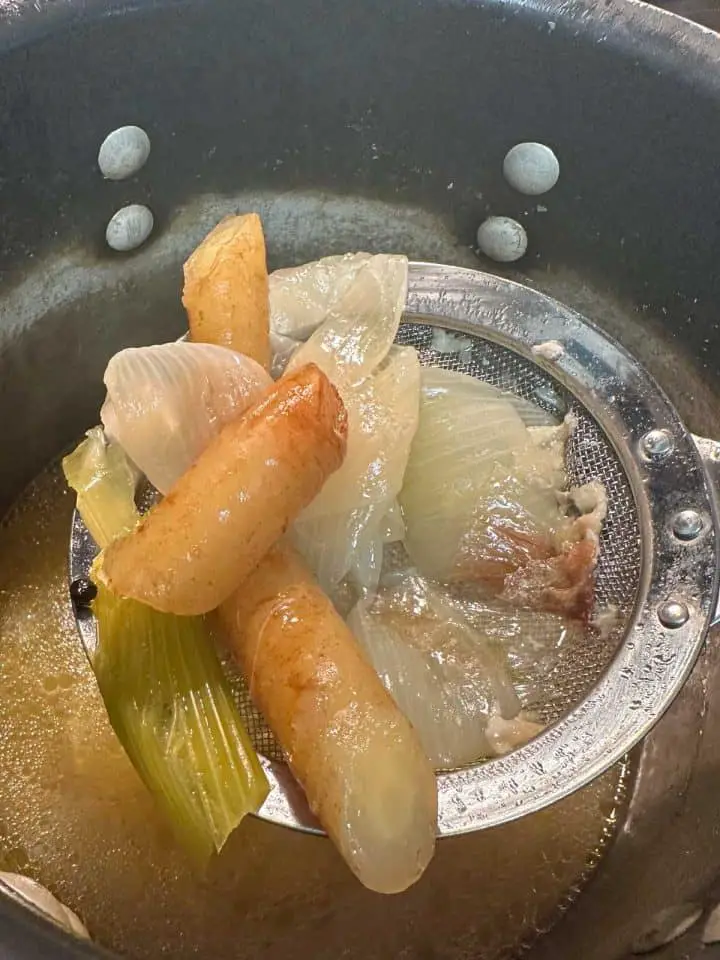 Homemade chicken stock in a large stockpot. There is a strainer spoon containing cooked celery, onions, and carrots that is closer to the camera and the large pot is in the background.