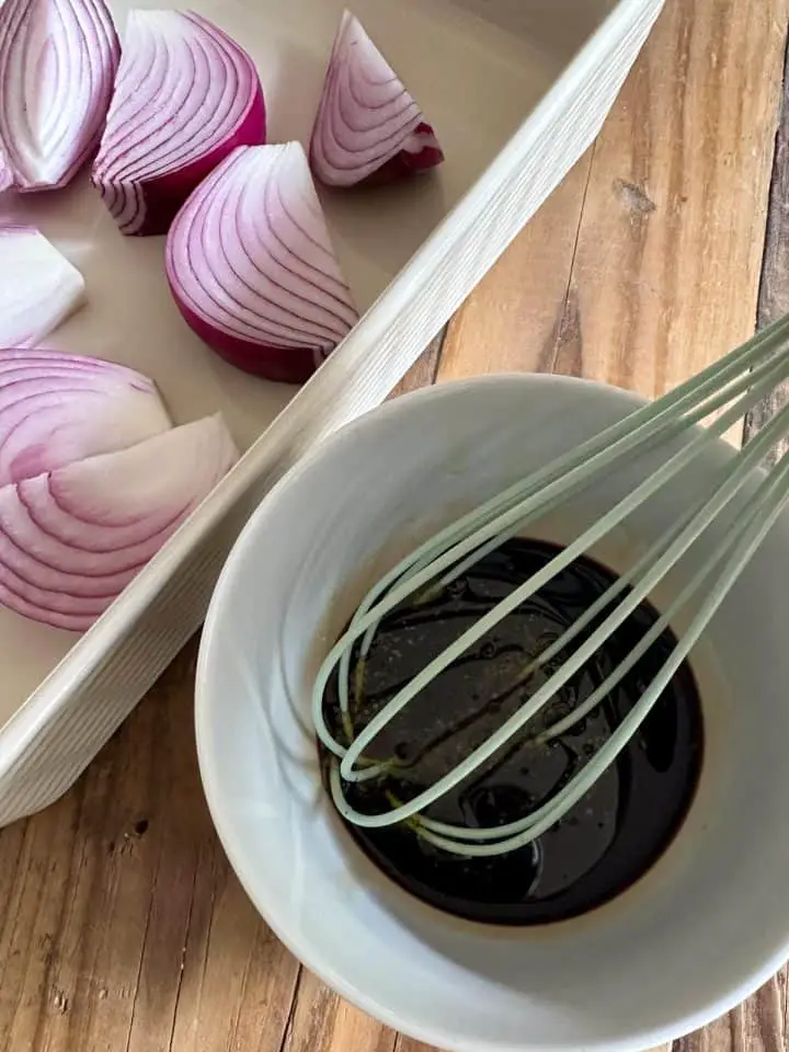 A casserole dish containing wedges of raw red onions, and a small bowl with balsamic vinegar with a whisk.