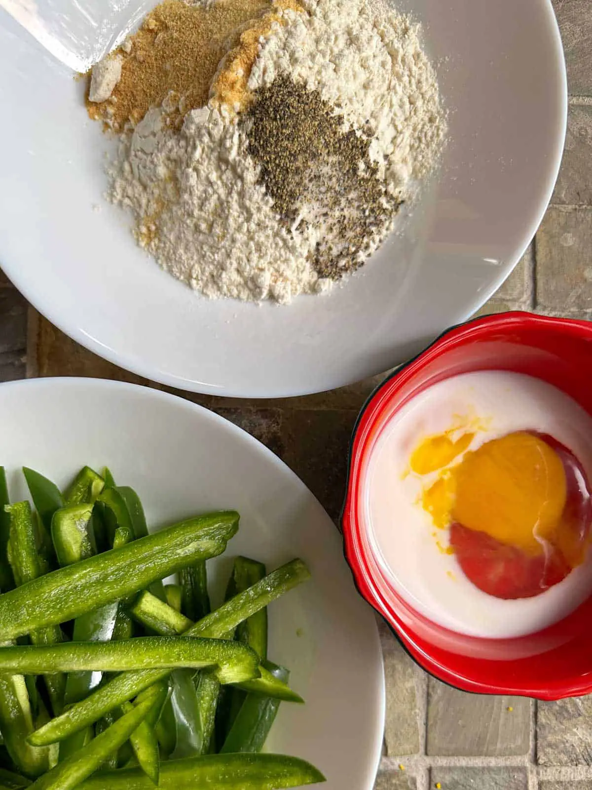 A white bowl containing flour and seasonings and a white bowl containing thin slices of jalapeños. There is also a small red bowl containing an egg and milk.