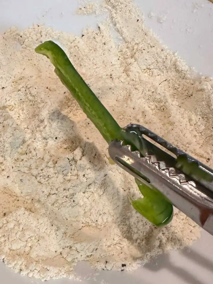 A bowl with seasoned flour and a pair of tongs holding a jalapeno slice above the seasoned flour.