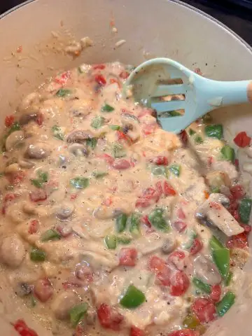 A Dutch oven containing chicken pieces, green bell pepper, and tomatoes in a creamy cheesy sauce. There is a blue slotted spoon resting in the Dutch oven.