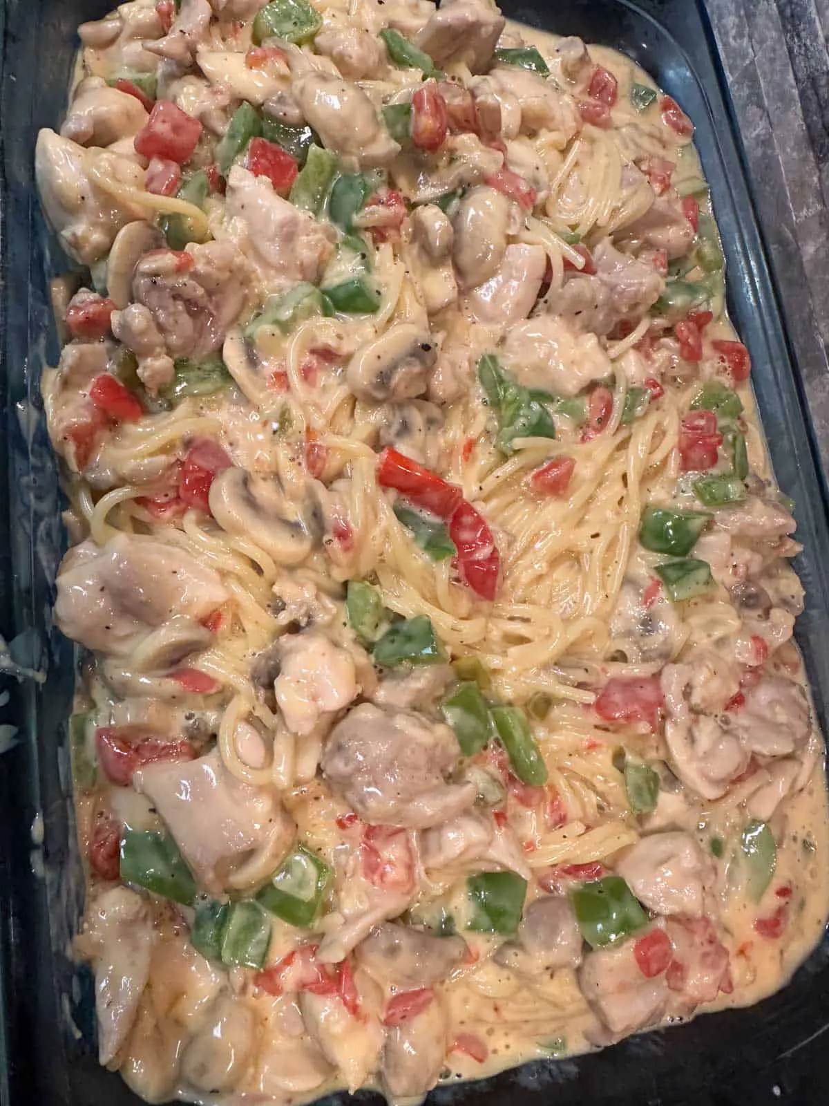 A glass casserole dish containing spaghetti, mushrooms, tomatoes, green bell peppers, and chilies in a creamy and cheesy sauce.