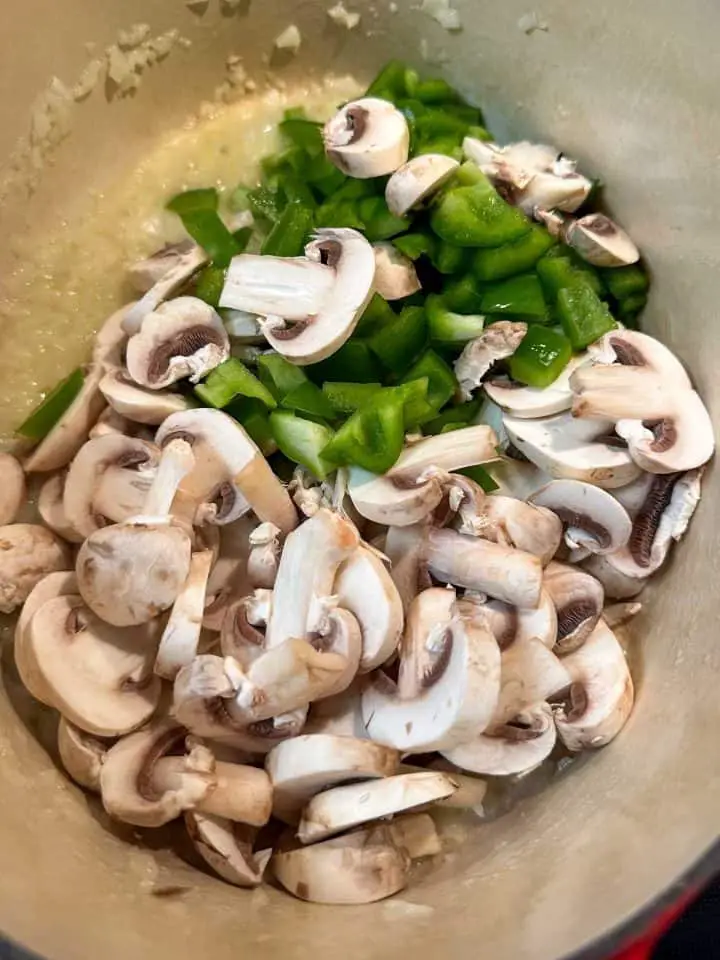 A Dutch oven containing sliced mushrooms, chopped green bell pepper, and minced onions.