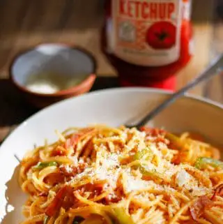 A white bowl containing Japanese Spaghetti Napolitan with sliced ham, sliced onions and sliced green bell pepper with grated Parmesan on top of the spaghetti. There is a fork resting in the bowl and a bottle of ketchup and small bowl with Parmesan in the background.