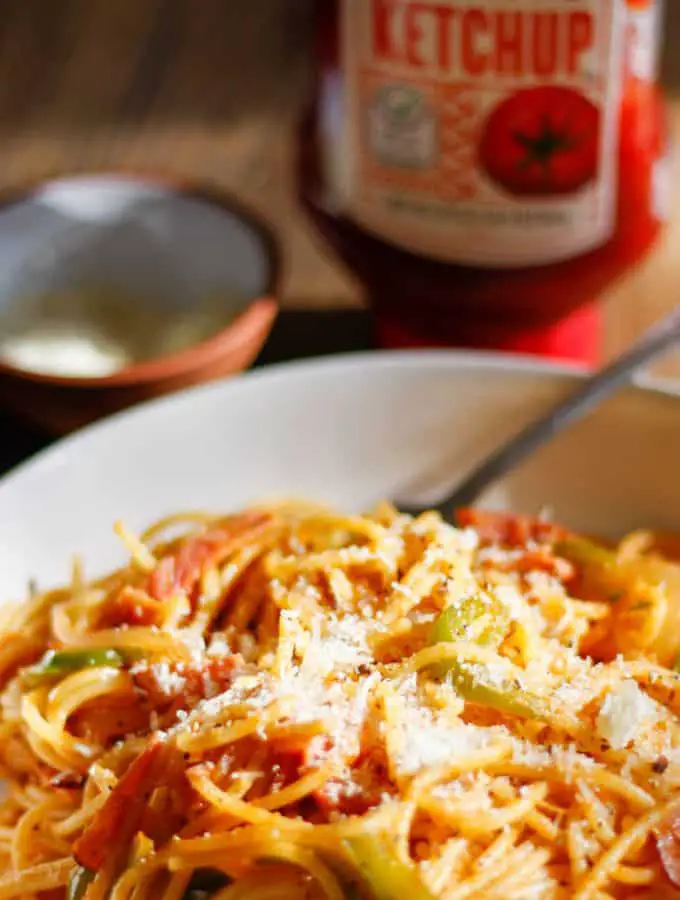 A white bowl containing Japanese Spaghetti Napolitan with sliced ham, sliced onions and sliced green bell pepper with grated Parmesan on top of the spaghetti. There is a fork resting in the bowl and a bottle of ketchup and small bowl with Parmesan in the background.