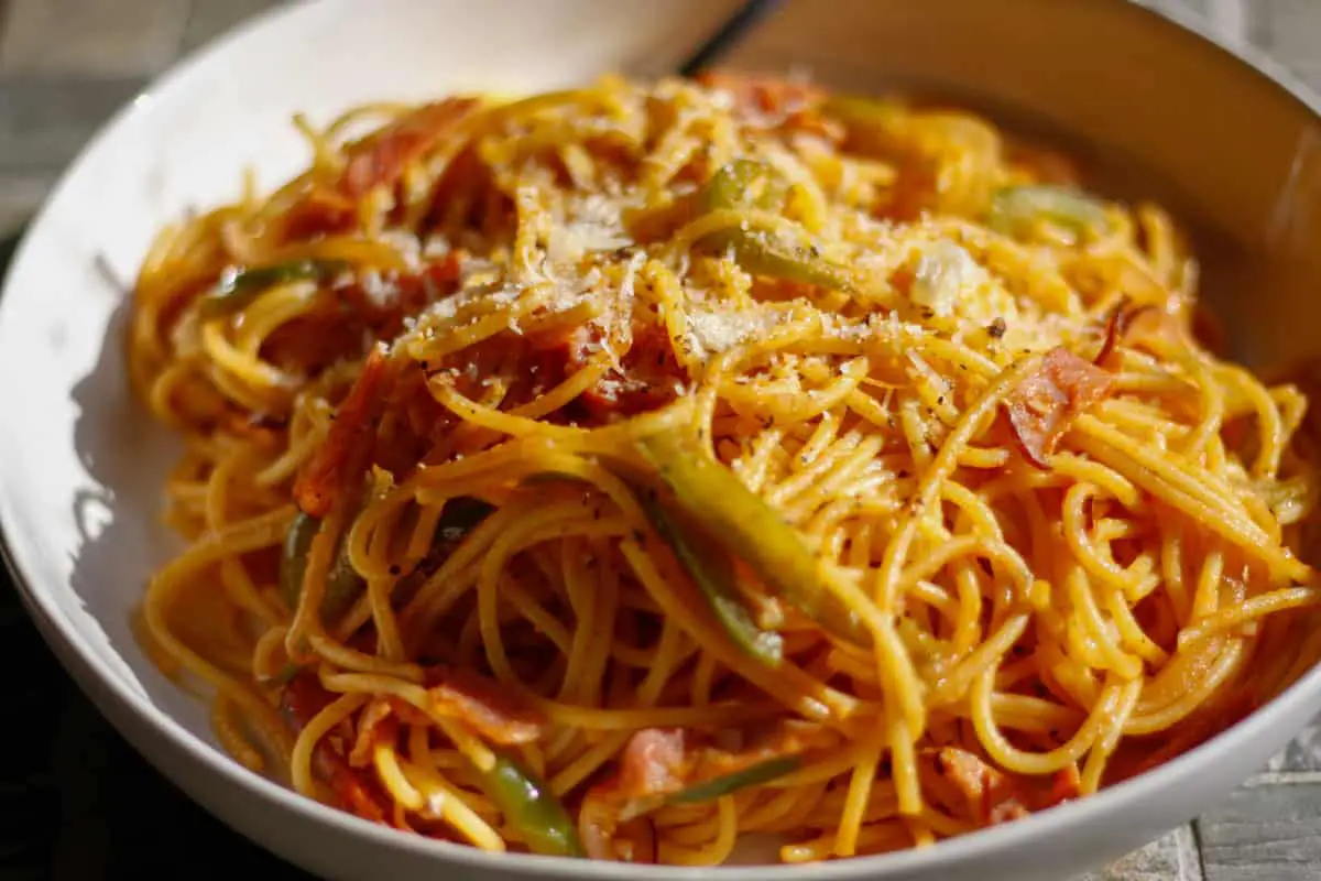 A white bowl containing Japanese Spaghetti Napolitan with sliced ham, sliced onions and sliced green bell pepper with grated Parmesan on top of the spaghetti. There is a fork resting in the bowl.