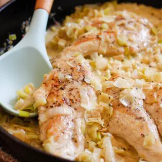 Chicken cooked with leeks in a cream sauce in a cast iron pan. There is a blue silicone spoon resting in the pan.