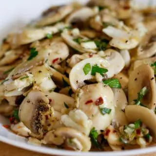 A white bowl containing sliced mushrooms with garlic, red pepper flakes, and minced Italian parsley.