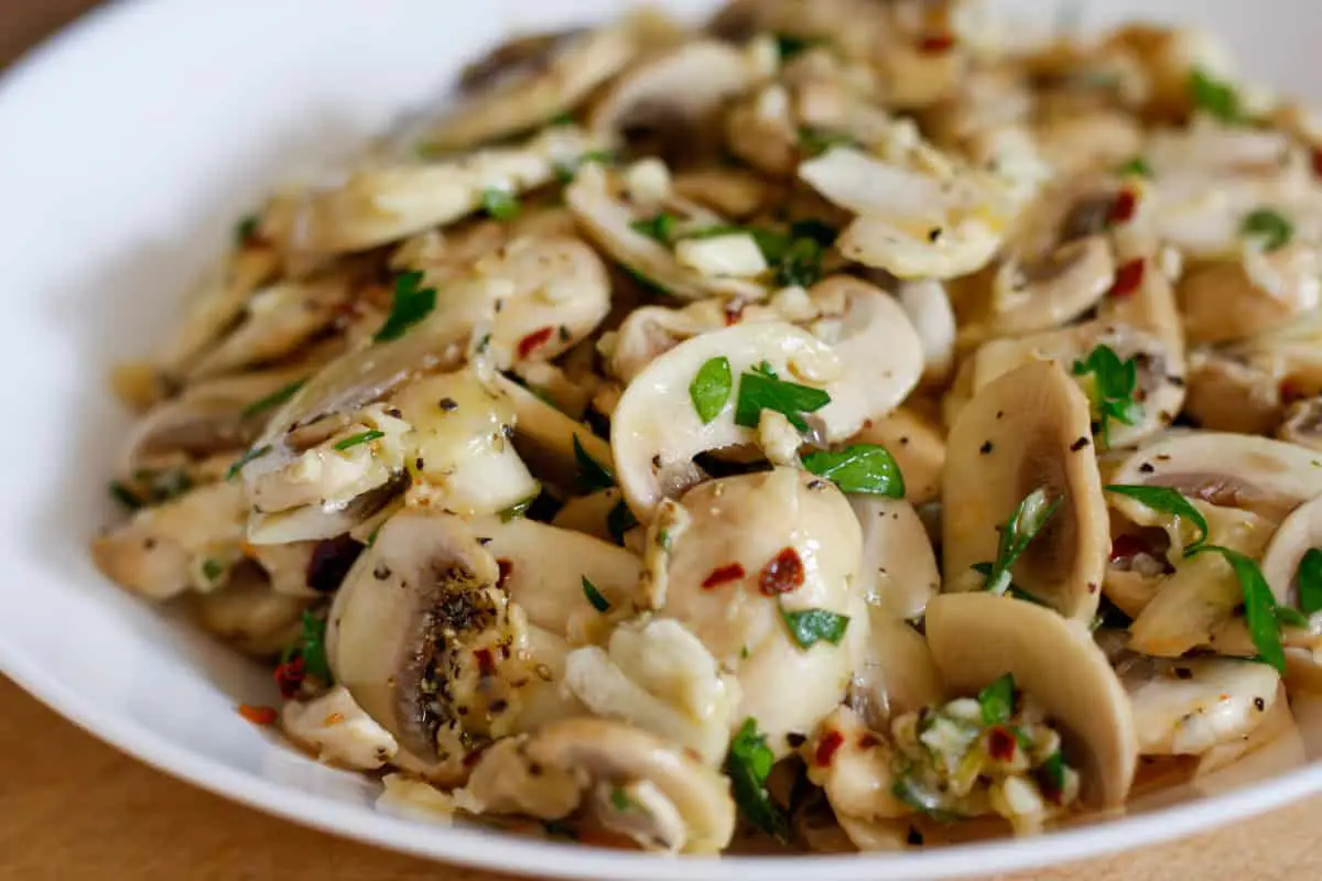 A white bowl containing sliced mushrooms with garlic, red pepper flakes, and minced Italian parsley.