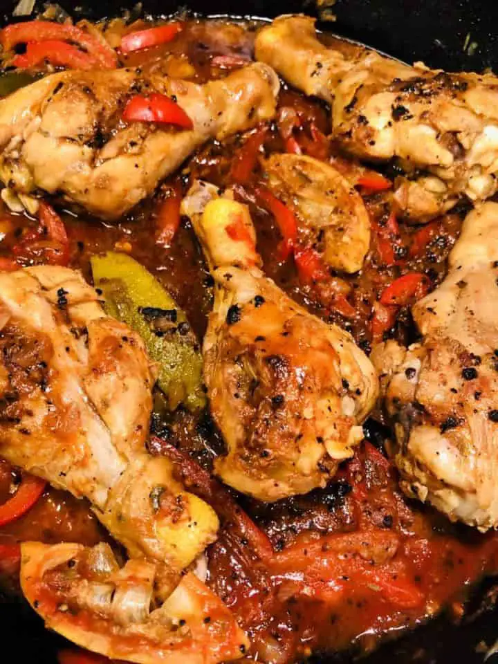 Haitian Chicken in a cast iron skillet in a savory red sauce with lime wedges and slices of red bell pepper.