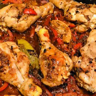 Haitian Chicken in a cast iron skillet in a savory red sauce with lime wedges and slices of red bell pepper.