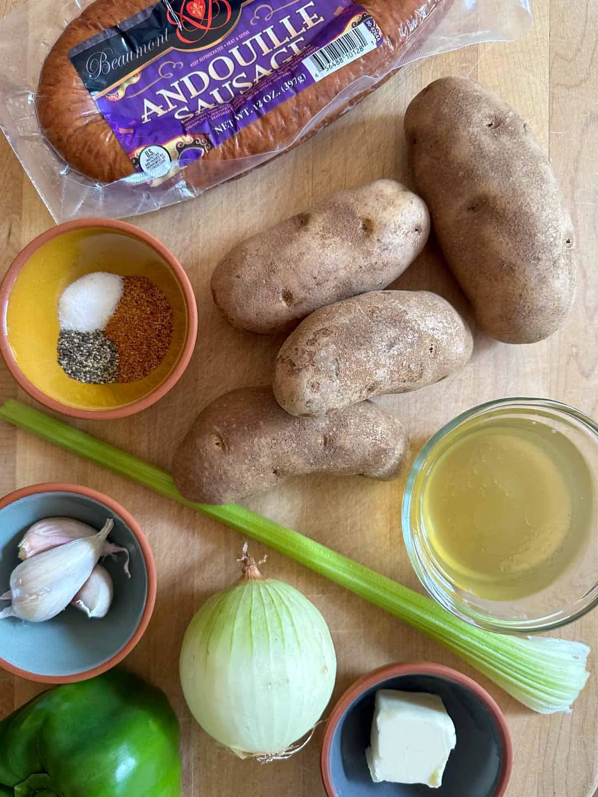 A package of Andouille Sausage, 4 russet potatoes, small bowls with seasonings, garlic, butter, and chicken broth. There is also a celery stalk, an onion, and a green bell pepper.