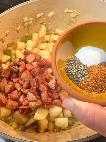 A Dutch oven containing small pieces of potatoes, onion, and green bell peppers along with small pieces of smoked sausage. There is someone holding a small bowl with seasonings above the Dutch oven.