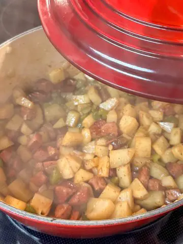 A red Dutch oven containing bite sized pieces of potatoes, green bell peppers and smoked sausage. There is steam coming from the Dutch oven and the lid of the Dutch oven is poised over the Dutch oven.