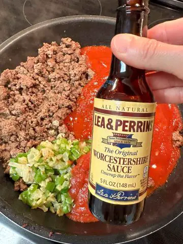 Sloppy joe sauce, tomato sauce, green bell pepper and onion, and ground beef in a large skillet. Someone is holding a bottle of Worcestershire Sauce in the foreground.