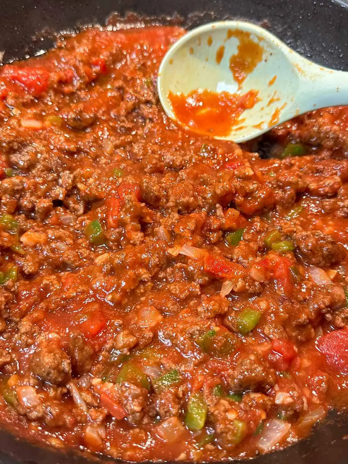 Sloppy joe sauce including beef, tomatoes, and green bell peppers in a skillet with a blue spoon resting in the skillet.