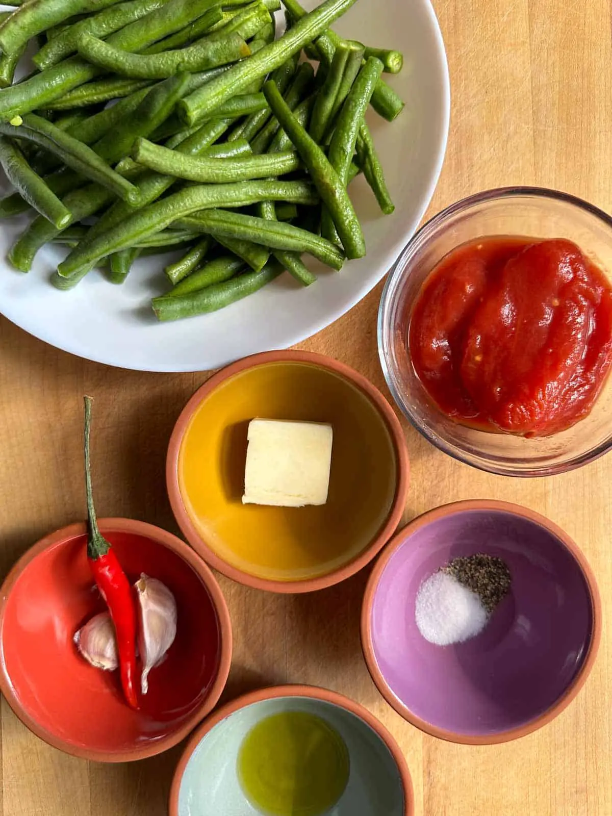 Green beans in a white bowl, and bowls containing Italian peeled tomatoes, butter, salt and pepper, olive oil, and garlic and chili.