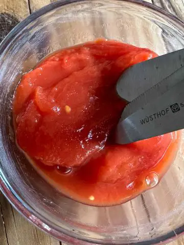 Italian peeled tomatoes in a glass bowl with a pair of Wusthof kitchen scissors poised over one of the tomatoes.