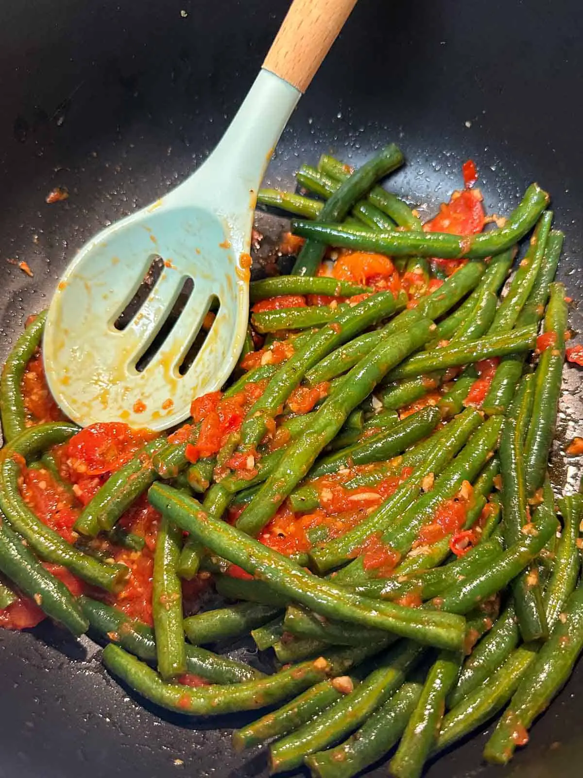 Green beans with tomatoes and garlic in a large pot. There is a blue slotted spoon with a wooden handle in the pot.