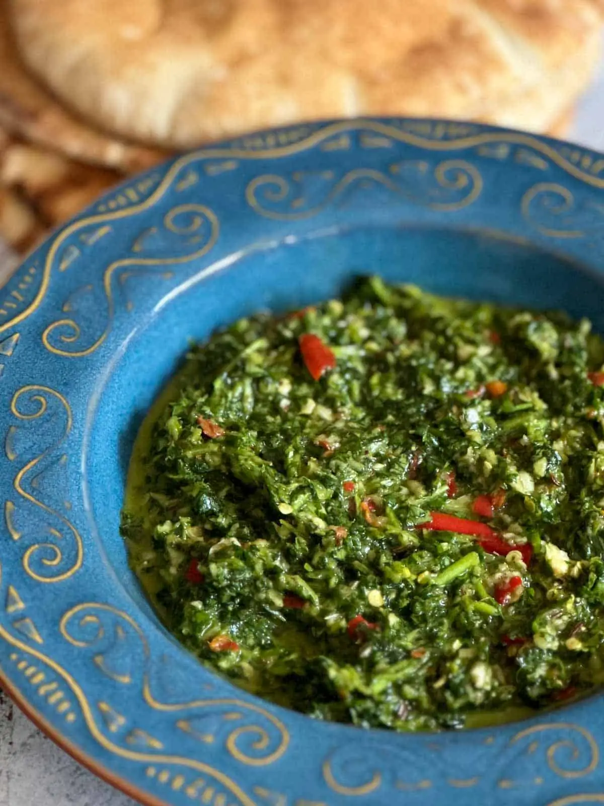 A blue bowl containing zhoug sauce which is blended cilantro with garlic, chilis, and seasonings. There are some pita breads in the background.