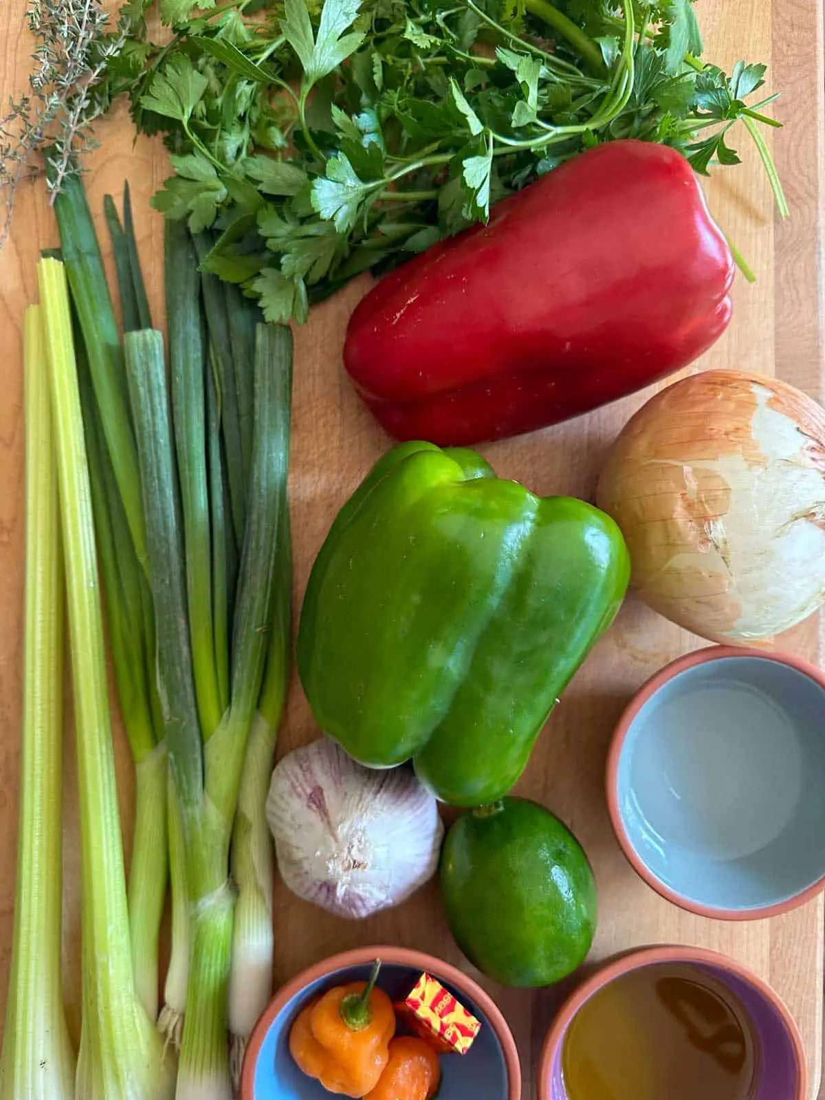 Thyme sprigs, parsley and cilantro, celery stalks, green onions, green and red bell pepper, onion, head of garlic, lime, bowls filled with olive oil and vinegar, and a bowl with Scotch bonnet chilis and a Maggi Bouillon cube.