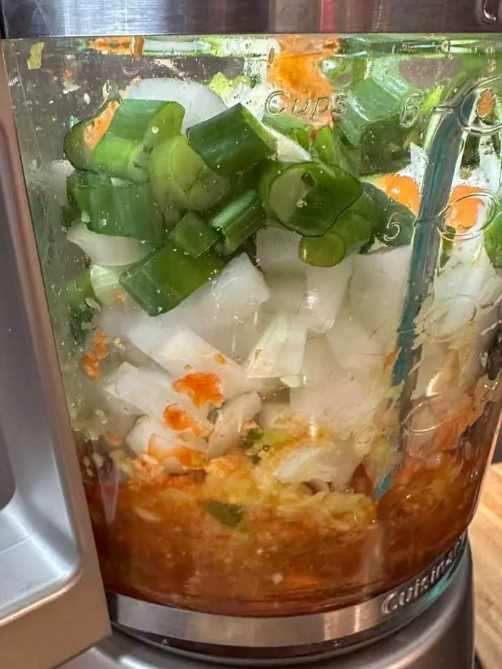 A blender containing blended vegetables and diced onions, chopped green onions, and scotch bonnet peppers.