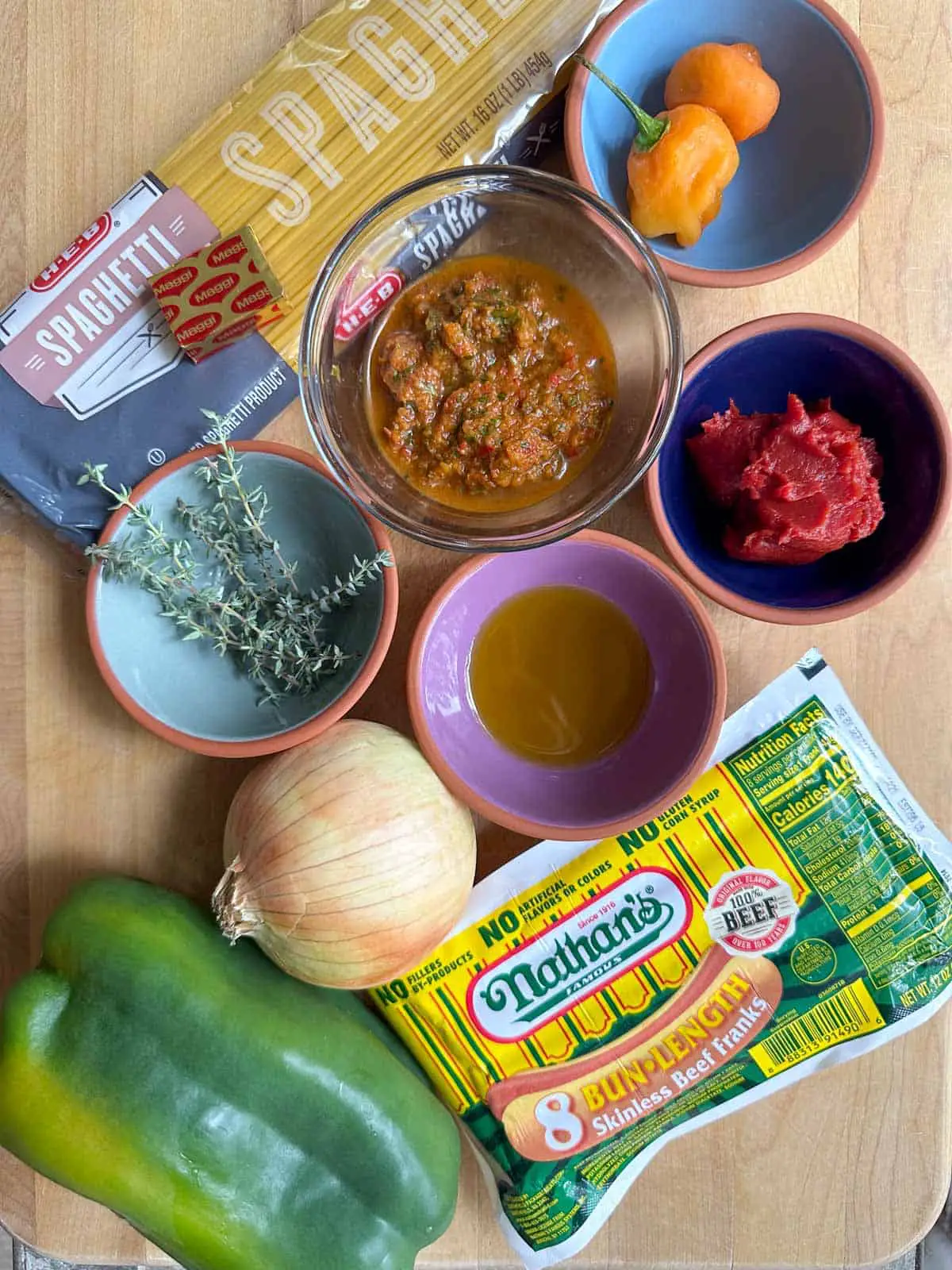 A package of spaghetti, Maggi chicken bouillon tablet, green bell pepper, onion, package of Nathan's hot dogs, and bowls containing Scotch bonnet chilis, Haitian epis, thyme, olive oil, and tomato paste.