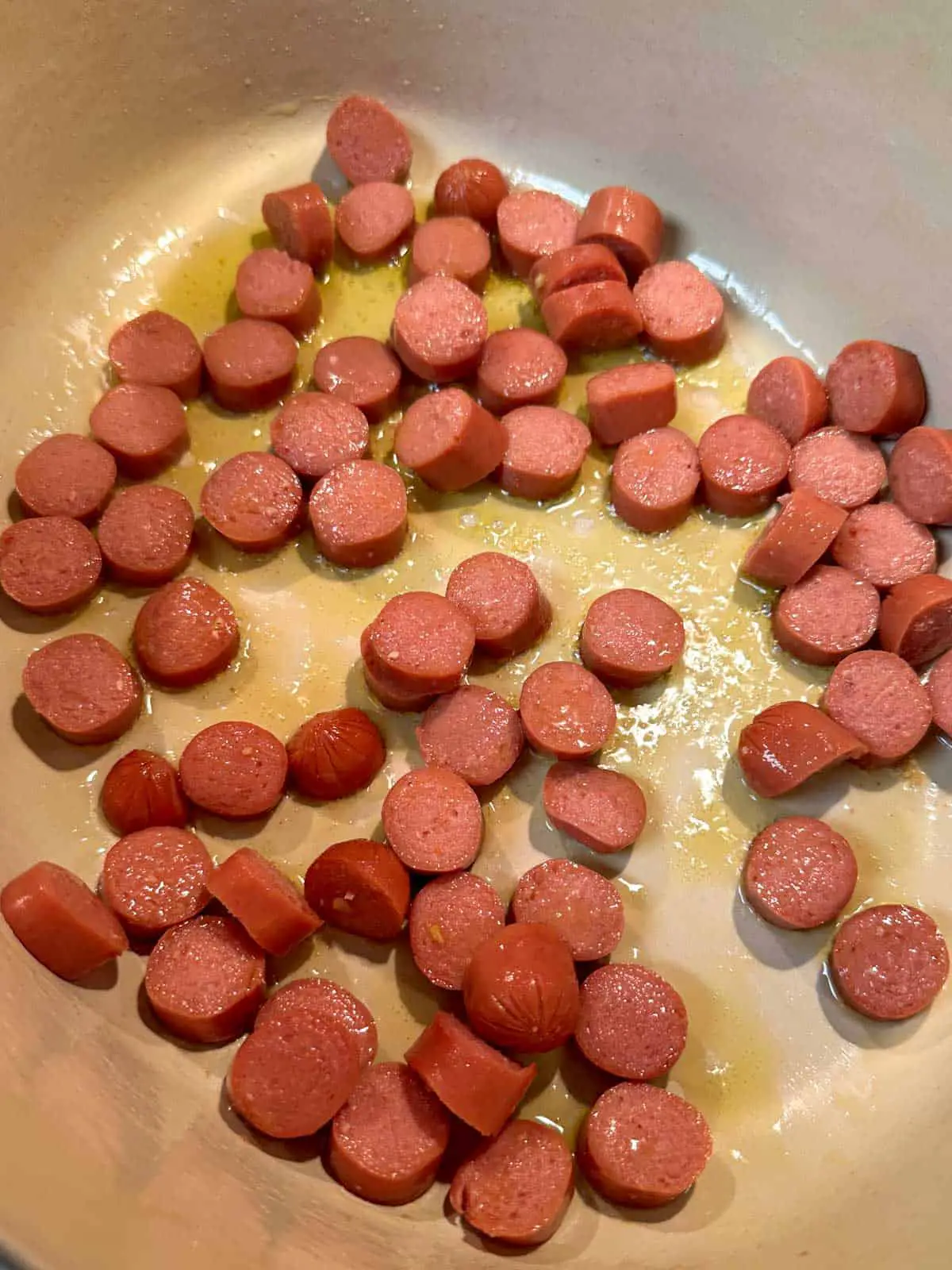 Slices of hot dogs cooking in oil in a Dutch oven.