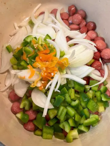 A Dutch oven containing sliced hot dogs, sliced onions, diced green bell pepper, and sliced Scotch bonnet chilis.