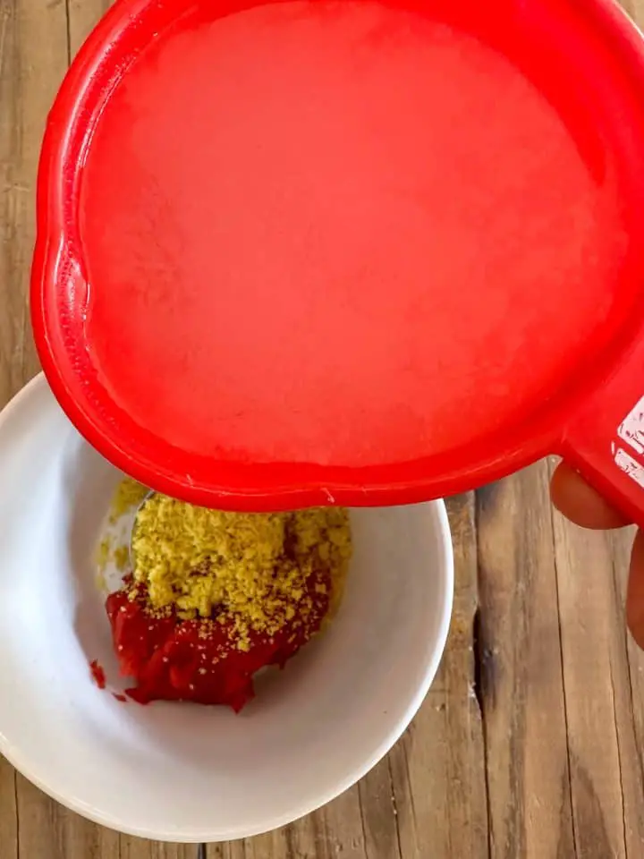 A white bowl containing tomato paste and crushed chicken bouillon cube with a red cup containing reserved pasta water poised over the bowl.