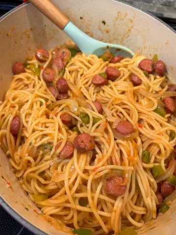 A Dutch oven containing Haitian spaghetti which is spaghetti with hot dogs, sliced onions, and diced green bell peppers. There is a blue spoon with a wooden handle resting in the Dutch oven.