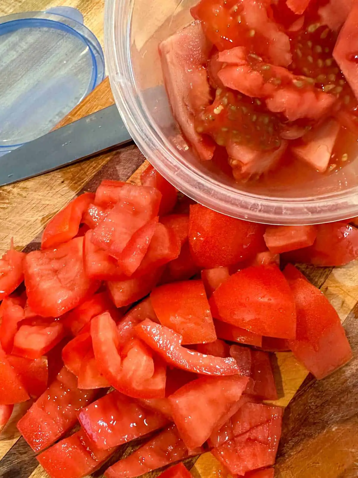 Diced tomatoes on a wooden cutting board. There is a small plastic bowl with the insides of the tomatoes in the bowl, and a blue lid for the bowl and a knife in the background.